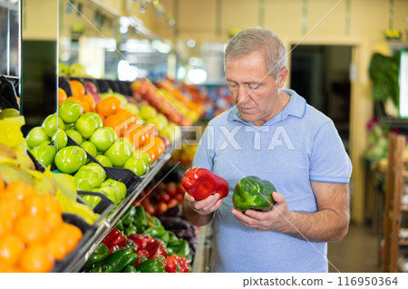 Focused elderly male pensioner in casual wear choosing local bell peppers during shopping in store 116950364