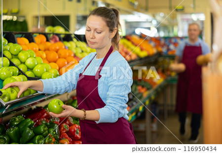 Middle-aged saleswoman putting apples on food stall in grocery store Middle-aged saleswoman putting apples on food stall in grocery store 116950434