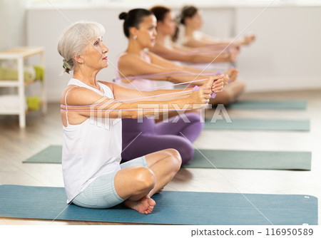 Women of different ages sit on mats and do pilates exercises with an expander in an gym Women of different ages sit on mats and do pilates exercises with an expander in an gym 116950589
