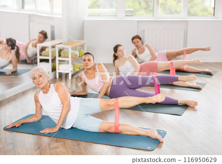Elderly woman lies on mat and performs stretching exercises with resistance bands in group pilates class 116950629