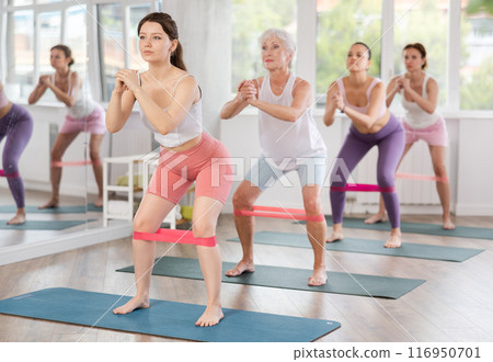 Group of women standing on mats and doing stretch exercises with resistance bands in gym 116950701