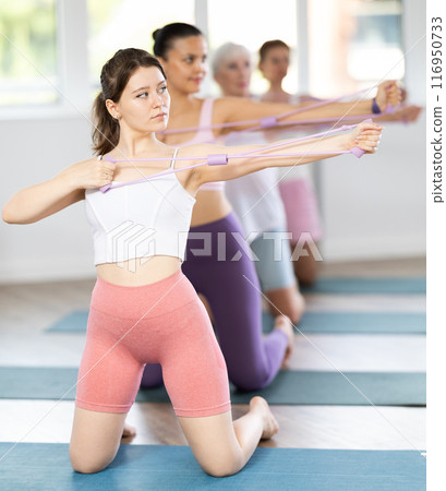 Group of women of different ages doing Pilates - kneeling and stretching their arms using an expander in gym 116950733