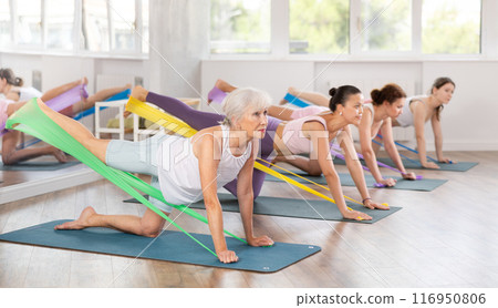Women on hands and knees doing stretch exercises for legs with resistance band on mats in gym 116950806