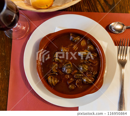 Flavored snails in shells in the tomato sauce in the white ceramic plate on the tablecloth presented in a cafe Flavored snails in shells in the tomato sauce in the white ceramic plate on the tablecloth presented in a cafe 116950864