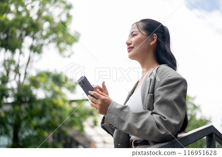 An attractive Asian businesswoman is standing outdoors, daydreaming and looking away. 116951628