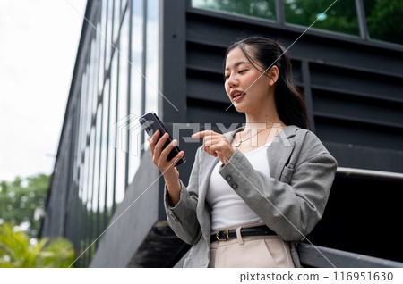 A beautiful Asian businesswoman is reading text or replying chat while standing outdoors. 116951630