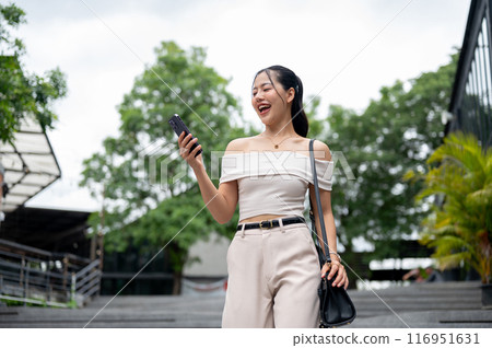 An attractive Asian woman is reading messages on her smartphone while walking in the city. An attractive Asian woman is reading messages on her smartphone while walking in the city. 116951631