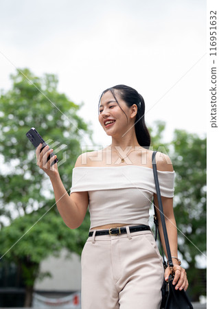 An attractive Asian woman is reading messages on her smartphone while walking in the city. An attractive Asian woman is reading messages on her smartphone while walking in the city. 116951632