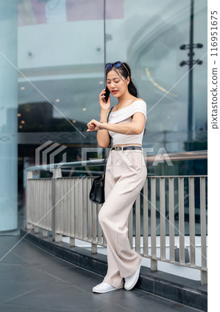 An elegant Asian woman is checking the time on her wristwatch while talking on the phone outdoors. 116951675