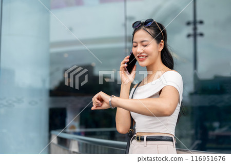 An elegant Asian woman is checking the time on her wristwatch while talking on the phone outdoors. 116951676