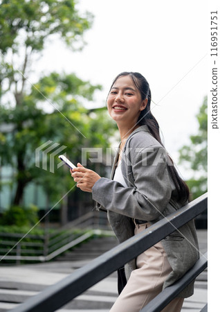 An attractive Asian businesswoman is standing outdoors in the city with a smartphone in her hand. 116951751