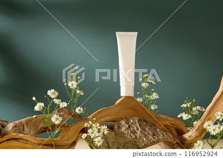 Frontal shot photography on dark blue background for advertising. A blank label cosmetic tube displayed on a log, with rock and tiny white flowers decorated around 116952924