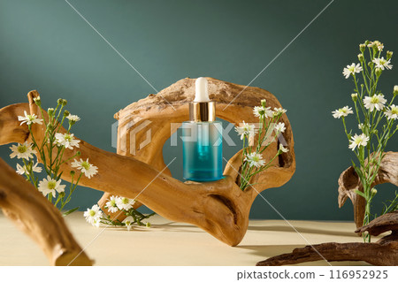 Front angle close up shot photography against dark blue background with natural theme. An unlabeled serum bottle is displayed on a crooked branch decorated with many clusters of tanacetum flowers Front angle close up shot photography against dark blue background with natural theme. An unlabeled serum bottle is displayed on a crooked branch decorated with many clusters of tanacetum flowers 116952925