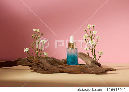 Blue serum bottle with gradient effect, displayed on the log with tanacetum flowers decorated on both sides, against on the pink background. Front view photo, blank space for advertising 116952941