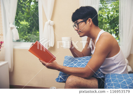 Young asian man reading book on cozy couch sofa at warm home. Relax man holding book read on sofa happiness. Man reading open book leisure mind in living room. Happy enjoyment man lifestyle at home 116953579