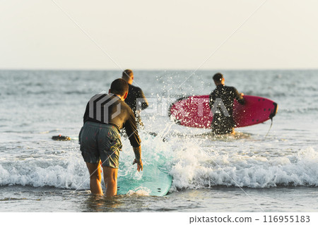 Three friends dressed in neoprene entering the sea with their surfboards Three friends dressed in neoprene entering the sea with their surfboards 116955183