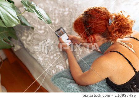 portrait of red-haired young woman lying on bed using mobile phone portrait of red-haired young woman lying on bed using mobile phone 116955459