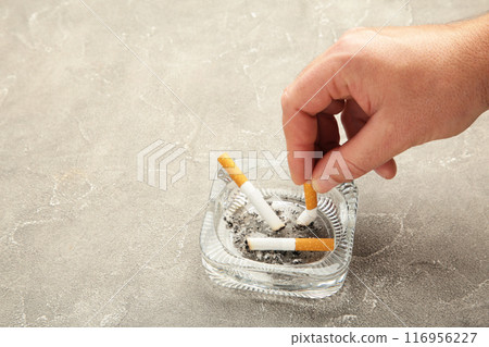 A hand puts out a cigarette in an ashtray on grey background. 116956227