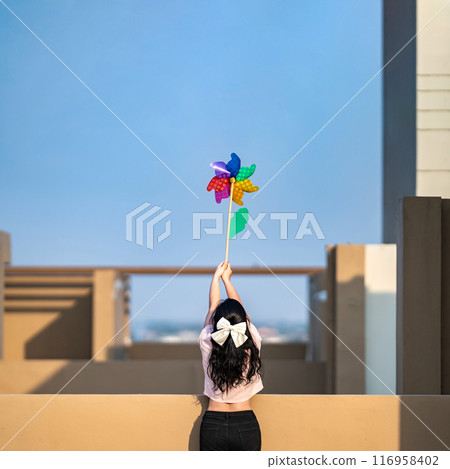 Asian woman posts and play Windmill Toy colourful rainbow colour on the building rooftop in sunset time. Asian woman posts and play Windmill Toy colourful rainbow colour on the building rooftop in sunset time. 116958402