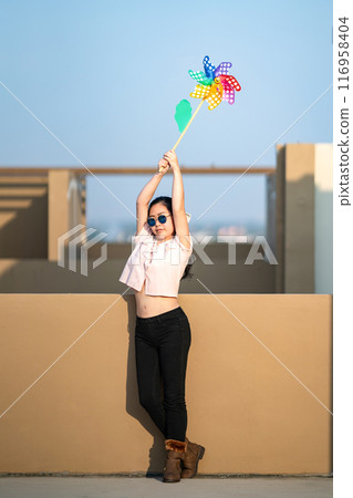 Asian woman posts and play Windmill Toy colourful rainbow colour on the building rooftop in sunset time. Asian woman posts and play Windmill Toy colourful rainbow colour on the building rooftop in sunset time. 116958404