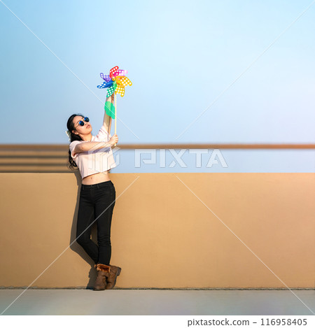 Asian woman posts and play Windmill Toy colourful rainbow colour on the building rooftop in sunset time. Asian woman posts and play Windmill Toy colourful rainbow colour on the building rooftop in sunset time. 116958405