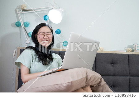 Glasses beautiful Asian woman is working on her laptop in the bedroom with a pastel green - brown color theme. Glasses beautiful Asian woman is working on her laptop in the bedroom with a pastel green - brown color theme. 116958410