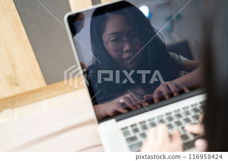 The reflection of Beautiful Asian woman from laptop screen. She plays laptop and lays on the bed in her bedroom. The reflection of Beautiful Asian woman from laptop screen. She plays laptop and lays on the bed in her bedroom. 116958424