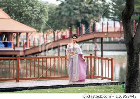A woman in a traditional Thai costumes is standing on the wooden bridge while being taken a portrait photo shooting. A woman in a traditional Thai costumes is standing on the wooden bridge while being taken a portrait photo shooting. 116958442