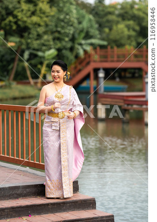A woman in a traditional Thai costumes is standing on the wooden bridge while being taken a portrait photo shooting. A woman in a traditional Thai costumes is standing on the wooden bridge while being taken a portrait photo shooting. 116958446