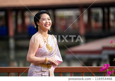A woman in a traditional Thai costumes is standing on the wooden bridge while being taken a portrait photo shooting. A woman in a traditional Thai costumes is standing on the wooden bridge while being taken a portrait photo shooting. 116958466