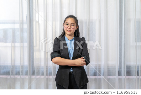 Little entrepreneur Asian eyeglasses woman with confident pose in black suit stands in front of window and curtain. 116958515