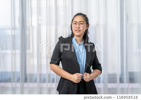 Little entrepreneur Asian eyeglasses woman with confident pose in black suit stands in front of window and curtain. 116958518