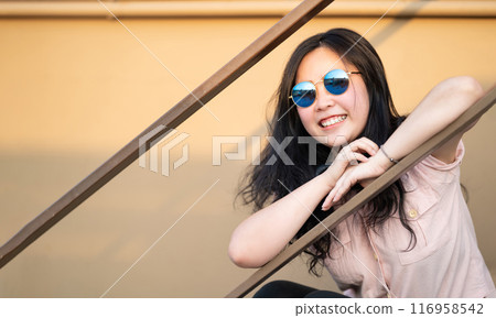 Asian happy woman is sitting on the stair and smile to camera in the sunset time at rooftop of the building. 116958542