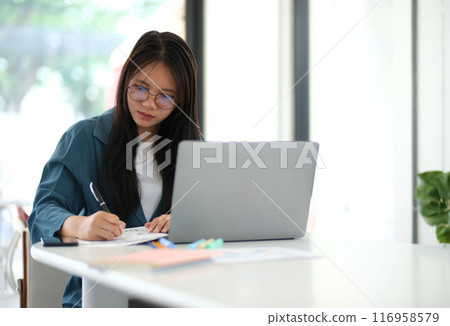 A woman is sitting at a desk with a laptop and a pen A woman is sitting at a desk with a laptop and a pen 116958579