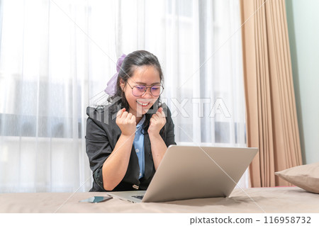 The woman was excited to look at a laptop which was laid on her bed because of her winning a lottery. She wearing glasses was screaming in her transparent curtain bedroom. The woman was excited to look at a laptop which was laid on her bed because of her winning a lottery. She wearing glasses was screaming in her transparent curtain bedroom. 116958732