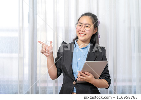 Happy Asian woman fingering up and pointing with tablet computer on her hand at copy space while standing in front of the transparent curtain see through the window light. 116958765
