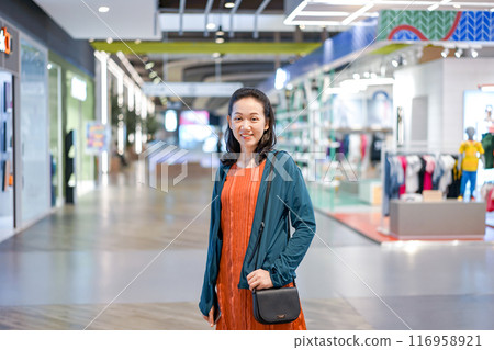 Woman wearing orange shirt and blue jacket put her hand in the pocket and smile with camera in shopping mall. 116958921