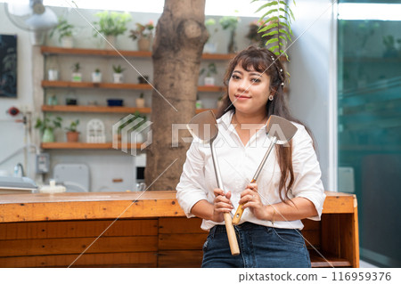 Asian woman wearing a white shirt and long jeans stands in the wooden counter kitchen holding two intertwined stove spades. 116959376