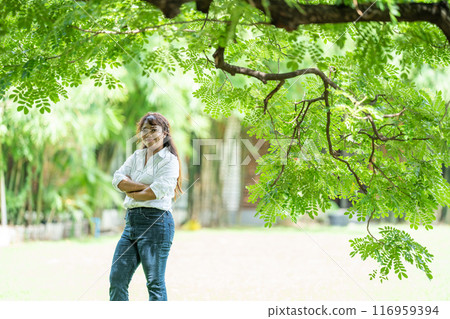Pretty woman wearing a trusty white jeans shirt and pretty smile to the camera under the tree outside field. 116959394