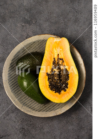 Two avocados and half a papaya on a clay plate made by hand on a gray background Two avocados and half a papaya on a clay plate made by hand on a gray background 116959489