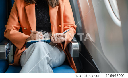 A woman in a bright orange blazer writing in a notebook while seated on an airplane, capturing a moment of productivity during travel. 116962001