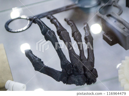 Art Object in form of human skeleton hand printed on 3D printer. Model prototype of skeleton printed on 3D printer from molten plastic on table. FDM 3D printing technology. New additive technologies Art Object in form of human skeleton hand printed on 3D printer. Model prototype of skeleton printed on 3D printer from molten plastic on table. FDM 3D printing technology. New additive technologies 116964479