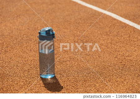 Blue refreshing bottle of water in sunny day with hard shadow on sports ground. Drinking water concept. Water balance for heat season for workout, activity and outdoor training. Close up. Copy space. 116964823