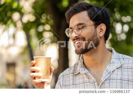 Happy Indian young man enjoying morning coffee hot drink and smiling standing on city street Happy Indian young man enjoying morning coffee hot drink and smiling standing on city street 116966137
