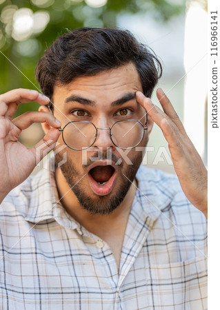 Young Indian man looking surprised at camera with big open eyes adjusting glasses on city street 116966141