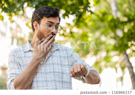 Serious dissatisfied young Indian man standing outdoors looking for someone waiting on city street 116966157