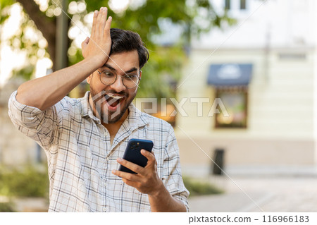 Hispanic young man using smartphone celebrating win good message news clenching fist on city street Hispanic young man using smartphone celebrating win good message news clenching fist on city street 116966183