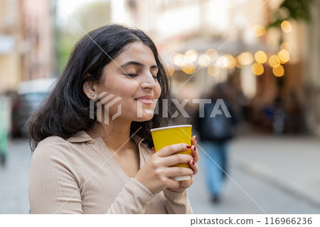 Happy Indian woman enjoying drinking morning coffee hot drink relaxing taking a break on city street Happy Indian woman enjoying drinking morning coffee hot drink relaxing taking a break on city street 116966236