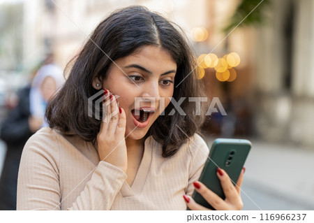 Excited happy winner Indian woman use smartphone celebrating win good message news on city street 116966237