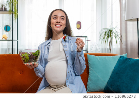 Happy pregnant woman with great appetite eats vegetable salad sitting on sofa healthy food self-care 116966278